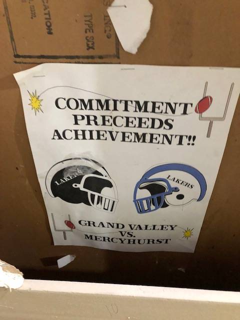 A sign on a box that reads, "Commitment Preceeds Achievement!" Grand Valley vs Mercyhurt. There are two helmets on the sign one that is black and the other that is blue and white. They both say "Lakers" on them.